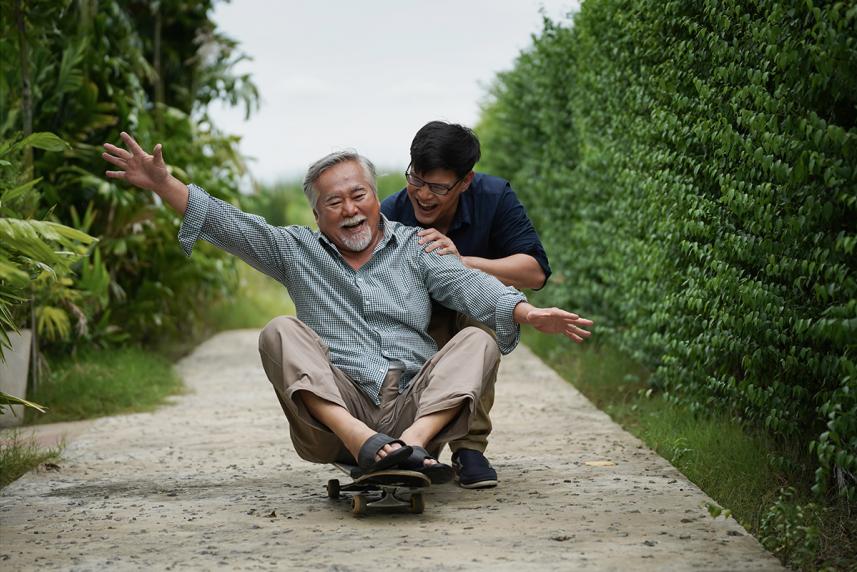 This photo shows an older man with gray hair and a beard sitting on a skateboard, laughing joyfully with his arms outstretched. A younger man with glasses and short black hair is behind him, holding onto his shoulders and smiling warmly. 